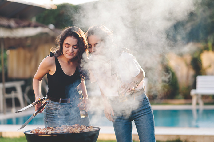 Kaksi nuorta naista on kumartunut pullot kädessään pallogrillin ääreen ihailemaan grillissä kypsyviä ruokia. Hiiligrillin savu ympäröi naiset ja taka-alalla näkyvän uima-altaan.